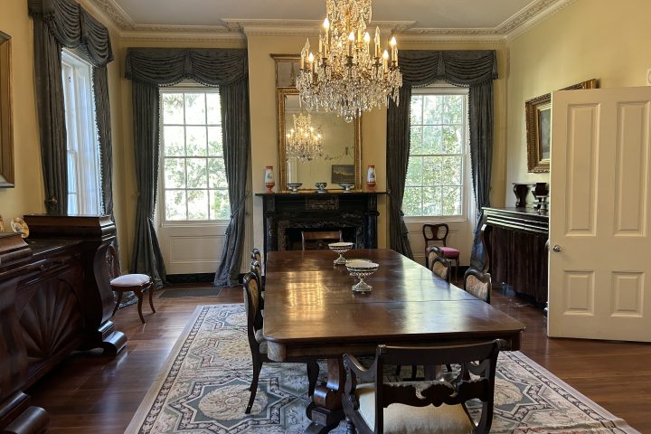 Elegant dining room with chandelier, long table, ornate chairs, and large windows with drapes.