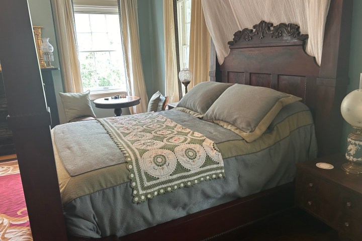 Elegant bedroom with a dark wooden bed, greenish bedding, crochet coverlet, and round side table near windows.