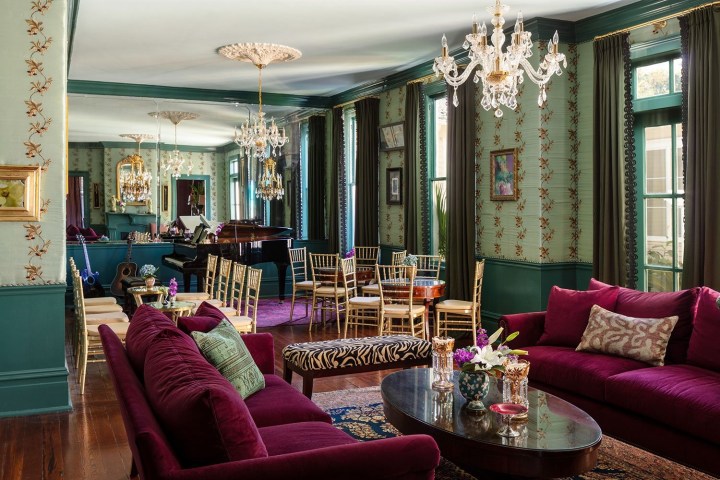 Elegant living room with chandeliers, burgundy sofas, patterned walls, and a grand piano.