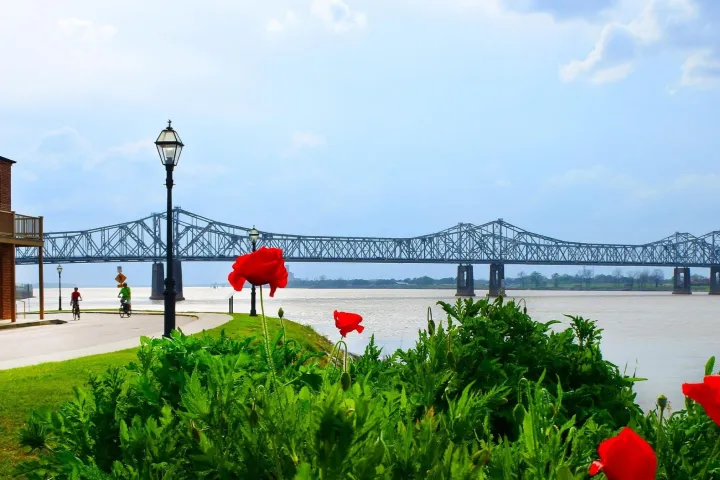 Steel bridge over river with brick building, red flowers, and cyclists in foreground.