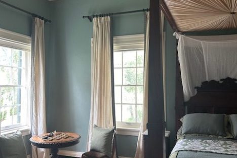 Elegant bedroom with canopy bed, chandelier, table, and two chairs by large windows.