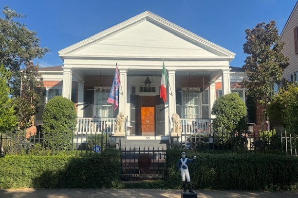 White house with porch, flags, and statues, surrounded by greenery and iron fence.