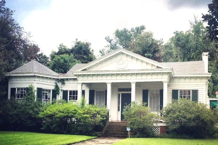 White, single-story house with pillars and green shutters surrounded by trees and shrubs.