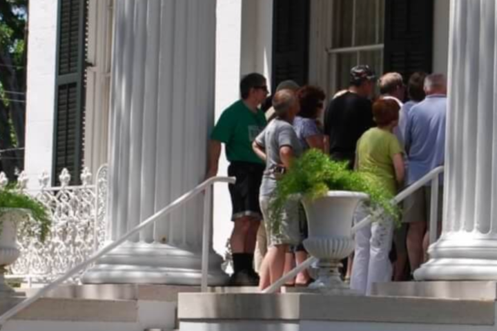 People gathered on steps of a white building with large columns and potted plants.