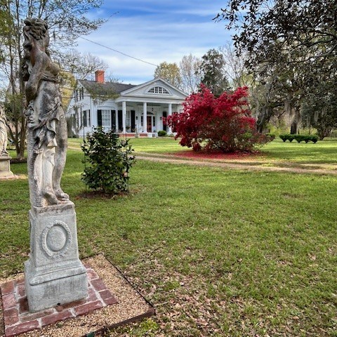 Stone statue in a garden with a historic building and red shrub in the background.