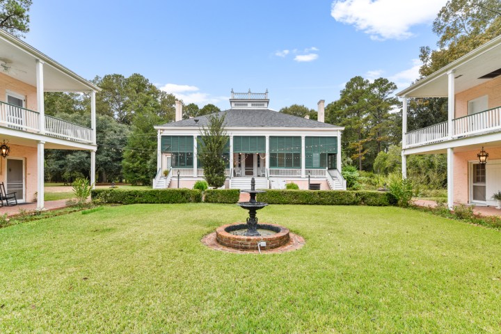 Historic mansion with white columns and green shutters, fronted by a fountain and landscaped grass.