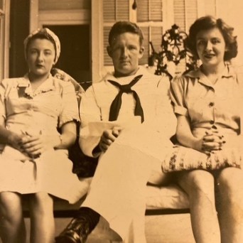 Three people seated on a bench, one man in a sailor uniform between two women in dresses.