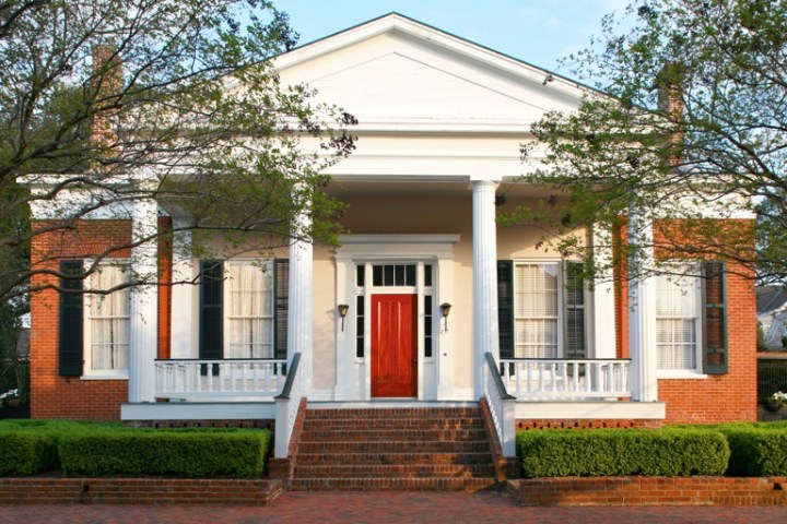 Historic red-brick house with white columns, a red door, and green shrubs.