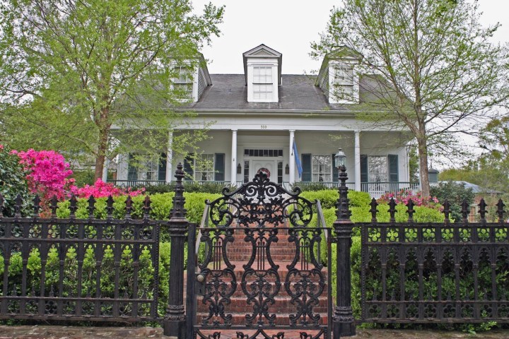 Two-story house with front porch, trees, flowers, and iron gate in the foreground.