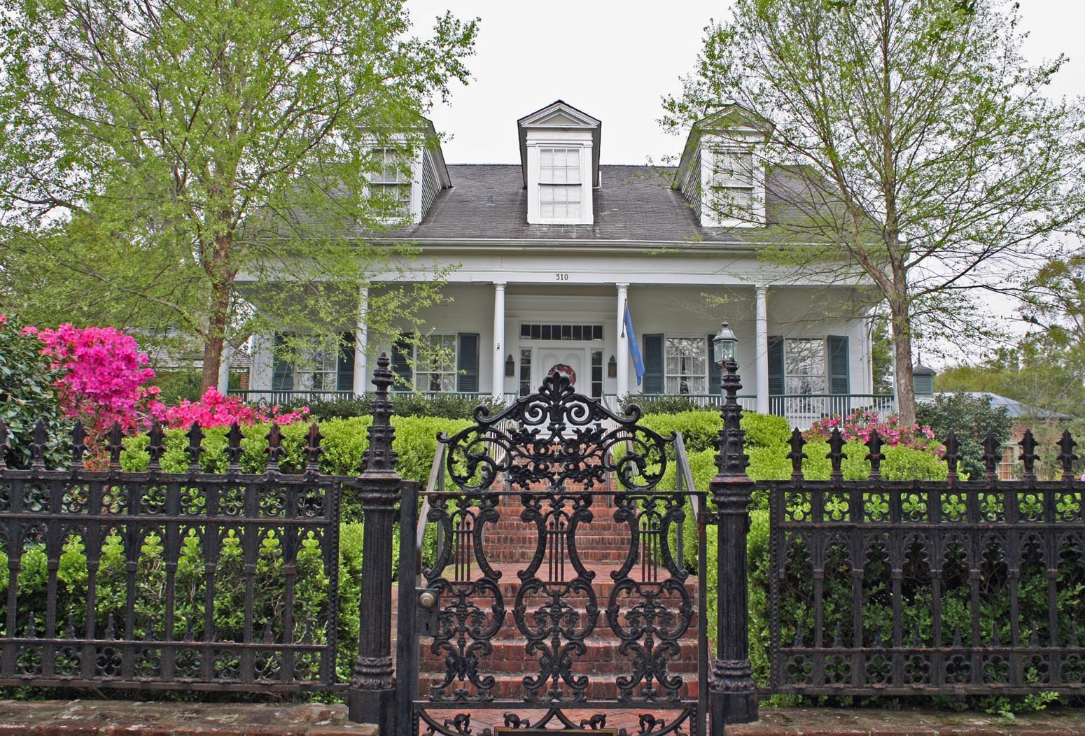 Two-story house with front porch, trees, flowers, and iron gate in the foreground.