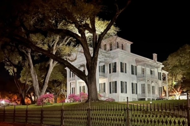 Illuminated white mansion with trees and a fence at night.