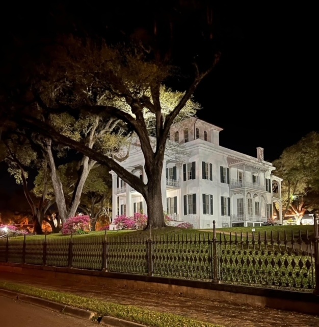 Illuminated white mansion with trees and a fence at night.